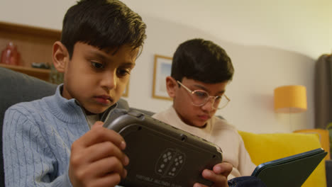 Dos-Niños-Sentados-En-Un-Sofá-En-Casa-Jugando-O-Transmitiendo-En-Una-Tableta-Digital-Y-Un-Dispositivo-De-Juegos-Portátil-3